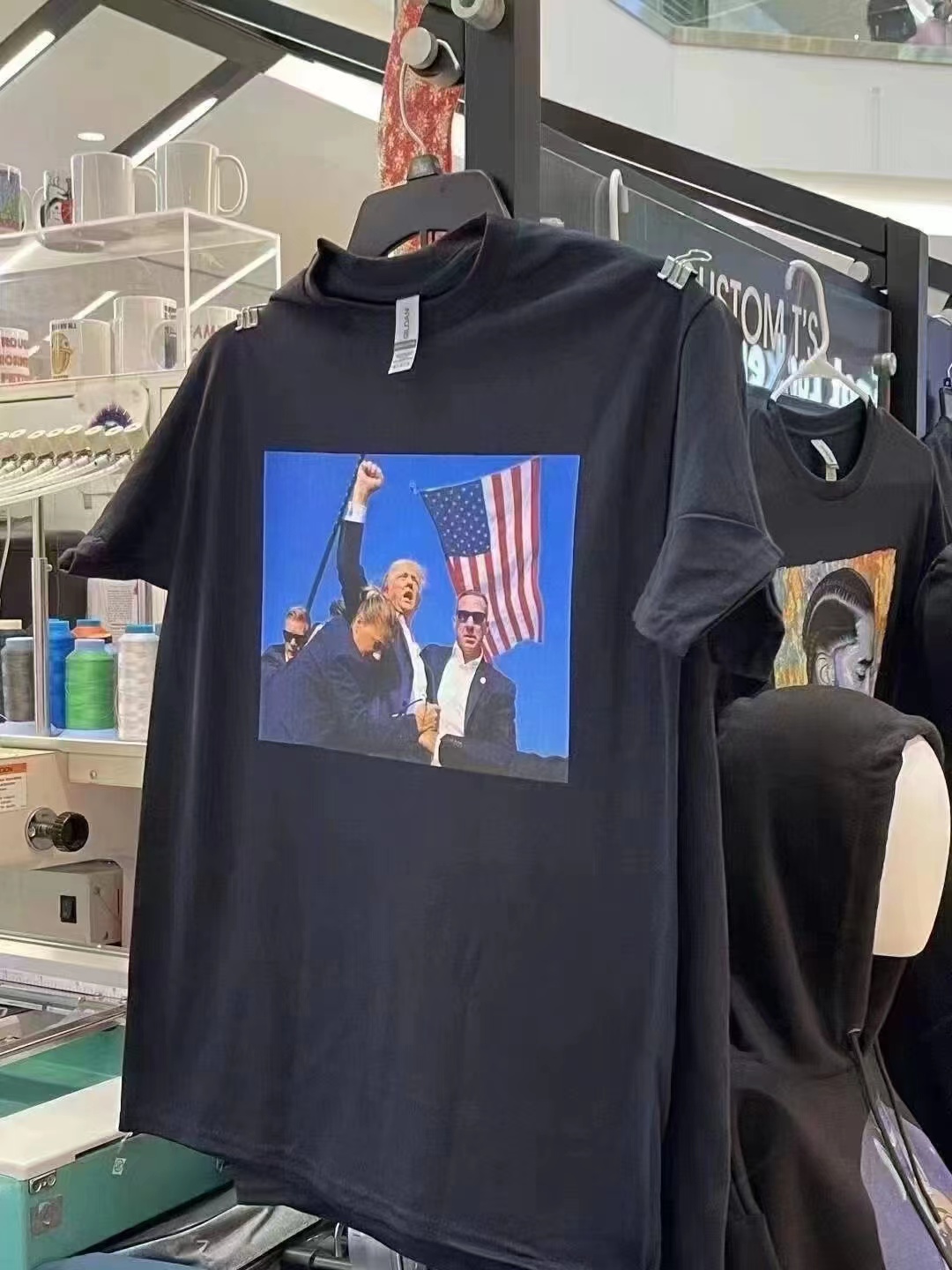 图片展示了一件挂在店内的黑色T恤，T恤正面印有三个人物图案，其中一人高举拳头，背景是美国国旗。T恤旁边还有其他衣物。图片位于介绍Zazzle平台内容的文档中，可能是用来说明在Zazzle上可以售卖带有创意图案的T恤等商品，为想在Zazzle上赚钱的小伙伴们提供参考。