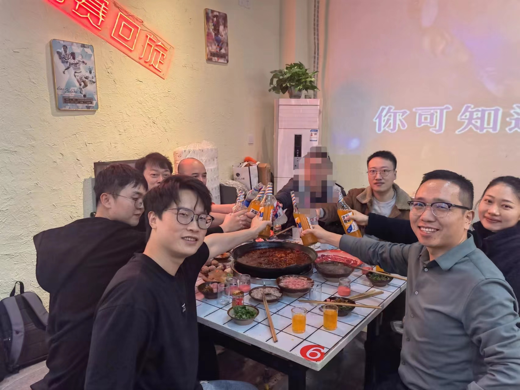 图片展示的是AI破局俱乐部重庆站线下聚会场景。画面中多人围坐在一张印有数字“6”的桌前，桌上摆放着火锅、饮料等食物。其中，左侧穿黑色上衣的男子和右侧穿灰色上衣的男子正举杯庆祝，其他人员也面带笑容。背景墙上挂着“你可知道”字样的装饰，还有植物和海报等装饰物。此图与文档中介绍的重庆站AI破局同城聚会内容相契合，呈现了聚会的欢乐氛围。