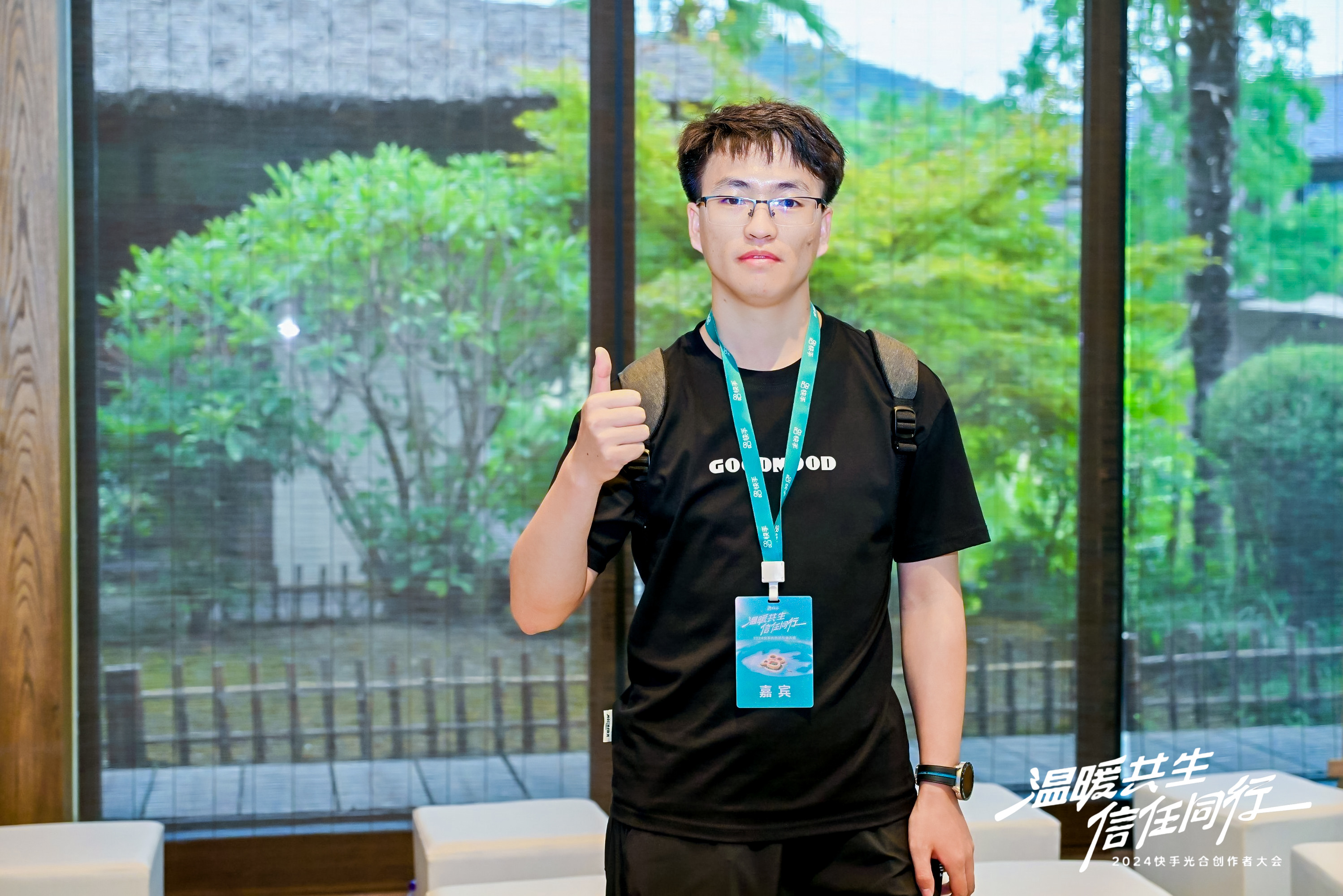 图片展示了一位身穿黑色T恤、佩戴蓝色证件的男子，他站在室内，背景是玻璃窗外的绿植和建筑。男子右手竖起大拇指，面带微笑，左手自然下垂。T恤上有“GOOD MOOD”字样。画面右下角有“2024快手光合开发者大会”的标识。此图可能是在2024快手光合开发者大会期间拍摄，男子可能是大会参与者或相关工作人员。