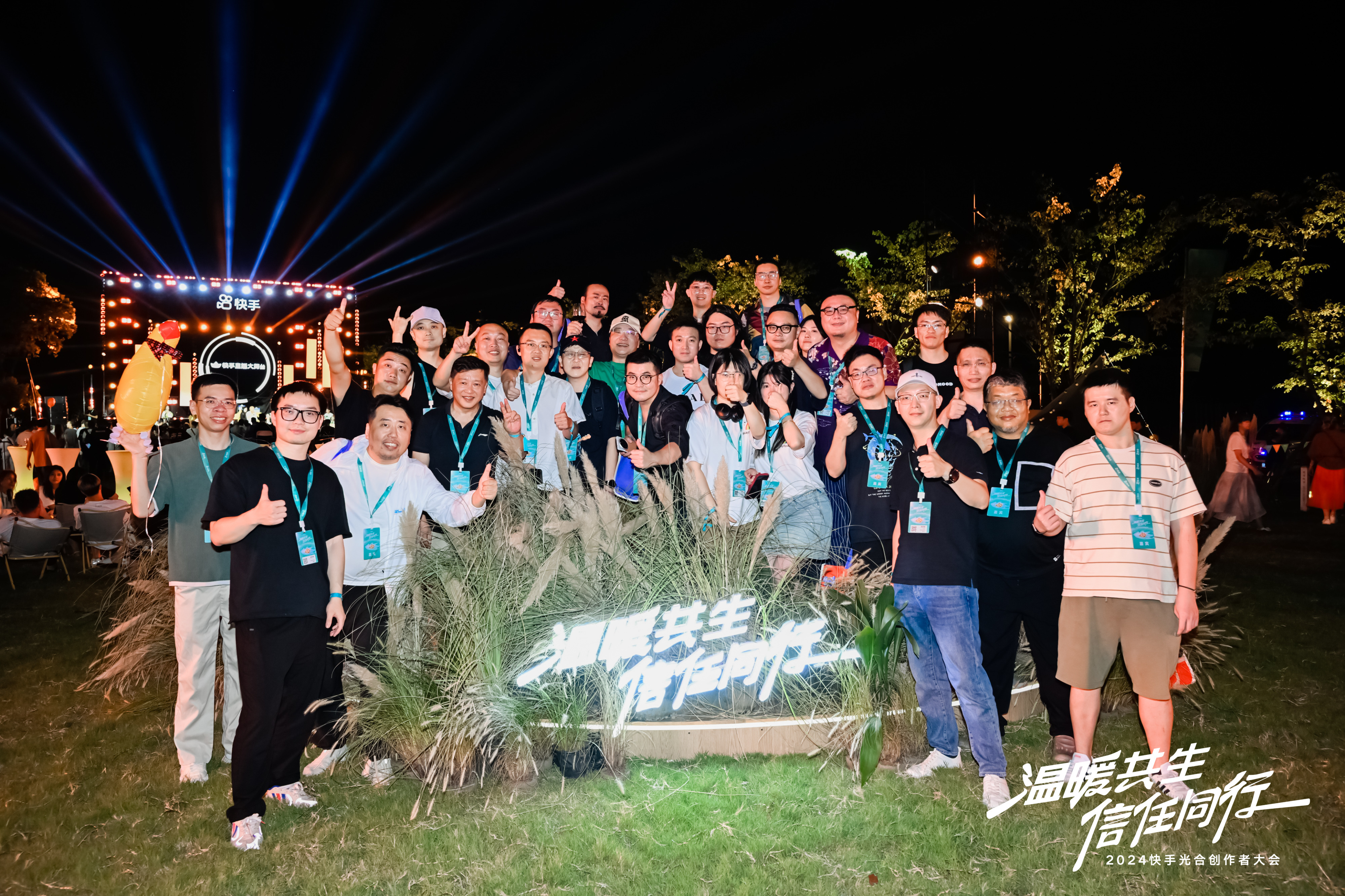图片展示的是2024快手光合开发者大会的参会人员合影。夜晚户外，众人站在“温暖共生 信任同行”字样的花丛前，面带笑容，有的竖起大拇指，有的比出胜利手势。背景有灯光装饰，远处可见舞台和观众席。图片与上文提到的2024快手光合开发者大会相呼应，记录了大会的欢乐时刻。