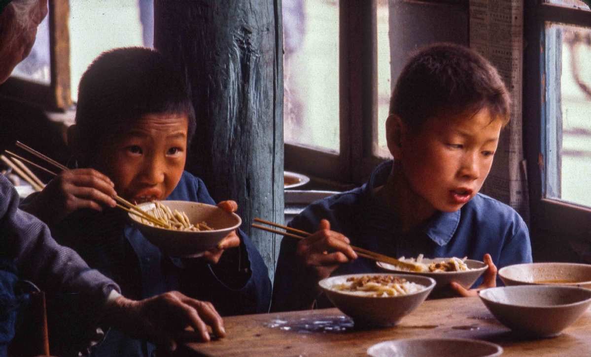 图片展示了两个小孩坐在桌前用筷子吃面的场景，桌上还有几个碗，背景中有一位老人和木质的窗户。此图位于“实操Part1：AI老照片转视频”文档中，在介绍电脑版【可灵】操作步骤，等待视频制作完成之后、提供视频下载链接之前，可能是使用【可灵】进行AI老照片转视频操作后生成视频中的画面内容示例。