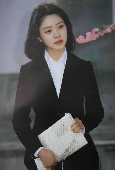 图片展示了一位身着黑色西装外套、内搭白色衬衣的女性，她手持一个白色手包，背景中有粉色花枝。此图位于介绍AI在老照片修复中可参与步骤的文档中，对应“图片高清化|美图秀秀”部分，其与上下文的关系是作为示例图片，分别展示旧的老照片以及使用美图秀秀【画质修复】功能高清放大之后的效果，直观呈现老照片修复中的图片高清化成果。