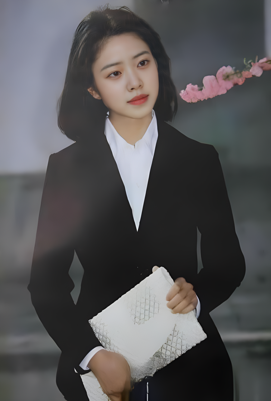 图片展示了一位身着黑色西装外套、内搭白色衬衫的女性，她手持白色手包，背景中有粉色花枝。这张图片位于介绍AI参与老照片修复过程中“图片高清化|美图秀秀”部分，是“旧的老照片”的示例图像，用于直观呈现老照片的原始状态，与后文“高清放大之后的老照片”形成对比，说明使用美图秀秀【画质修复】功能前后的变化情况。