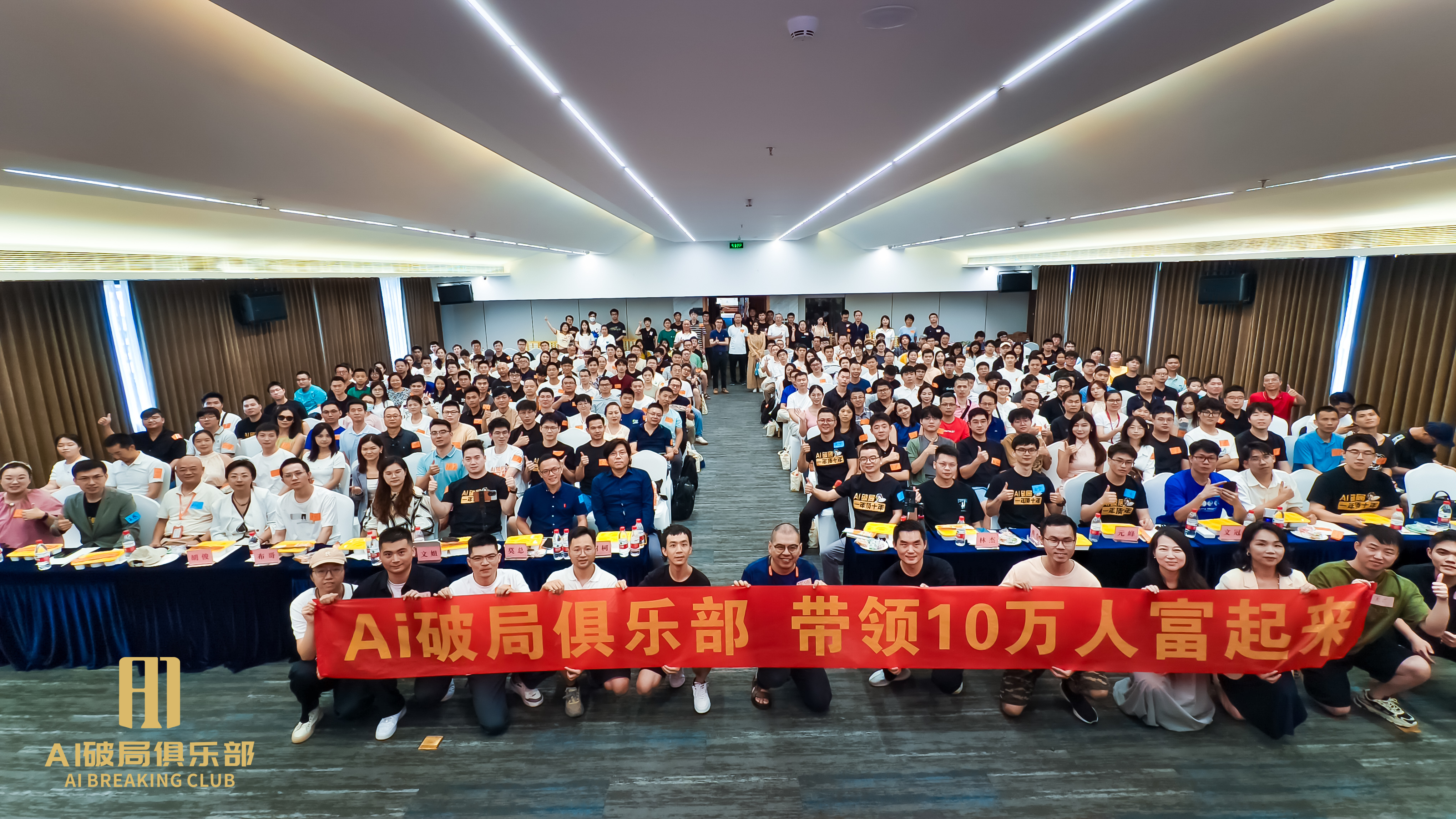 图片展示的是“AI破局俱乐部”线下大会场景。会场内坐满了人,大家围坐在长桌旁,桌上摆放着黄色的餐盘。画面中央有红色横幅,上面写着“AI破局俱乐部 带领10万人富起来”。会场背景为深色窗帘,两侧有蓝色桌布的长桌。图片与上下文紧密相关,上下文提到作者去年2月进入AI圈子后,上个月与文冠、树科一同参加了深圳场的线下大会,此图正是此次大会的现场照片。