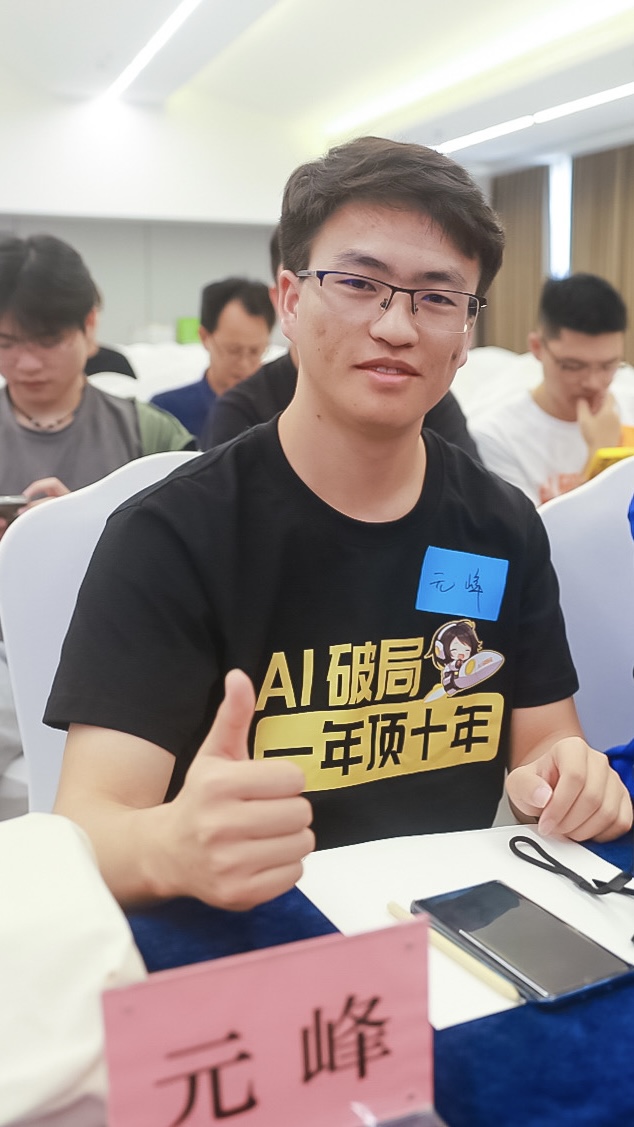 图片展示了一位穿着印有“AI破局 一年顶十年”字样的黑色T恤的男子，他坐在桌前，左手竖起大拇指，面带微笑，桌上放有手机等物品，桌前有写着“元峰”字样的粉色牌子。背景中还有其他人在使用手机。这张图片与文档中“破局年度拉新|小林战队”部分内容相关，可能是对文中提到的“小林战队”成员的展示，体现了团队成员的精神风貌。