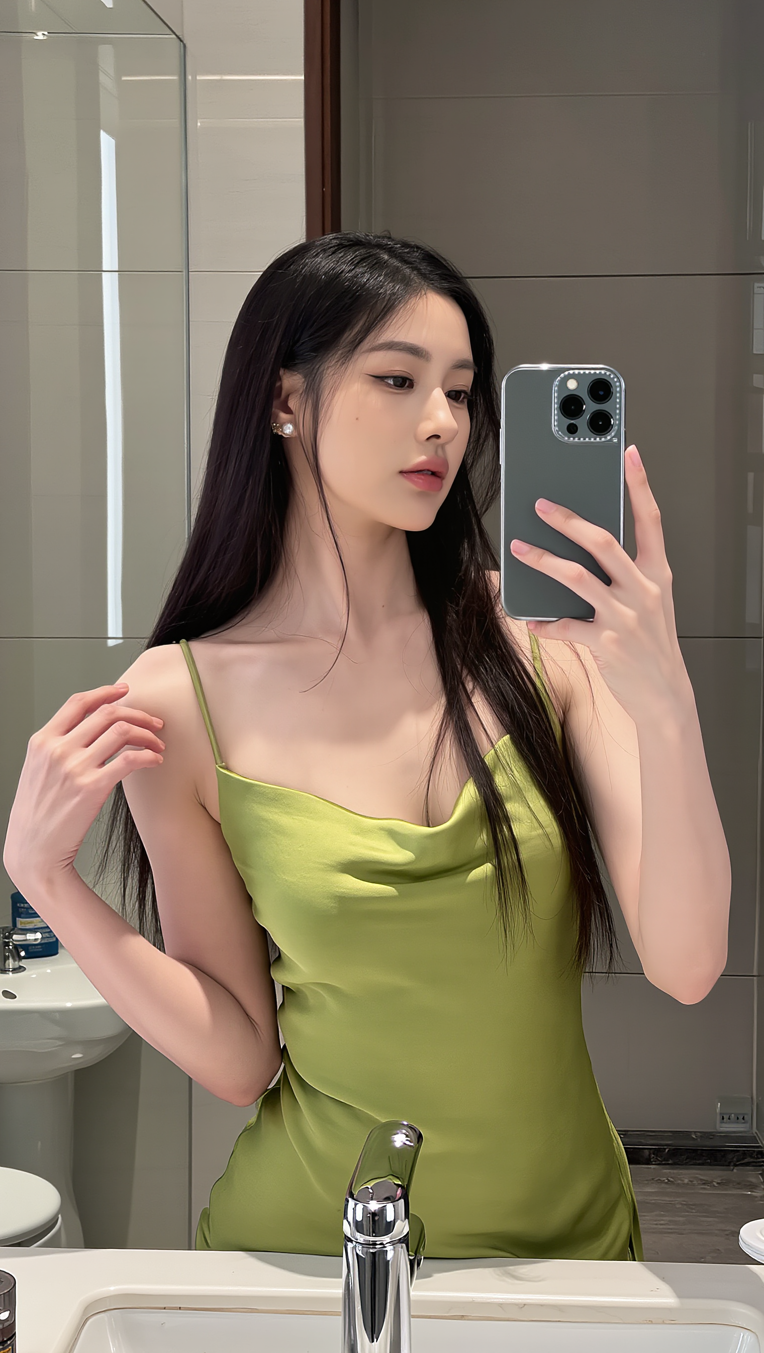 图片展示的是一位身着绿色吊带裙的长发女子在镜前自拍的场景。她手持一部后置摄像头有多个镜头的手机，背景为带有洗手池、镜子等设施的室内环境。此图位于介绍一键将图片转换为完美JSON、实现AI图像生成100%还原原图的文档中，在“复刻后的图片”相关内容处，是用于展示AI图像生成复刻效果的案例原图。