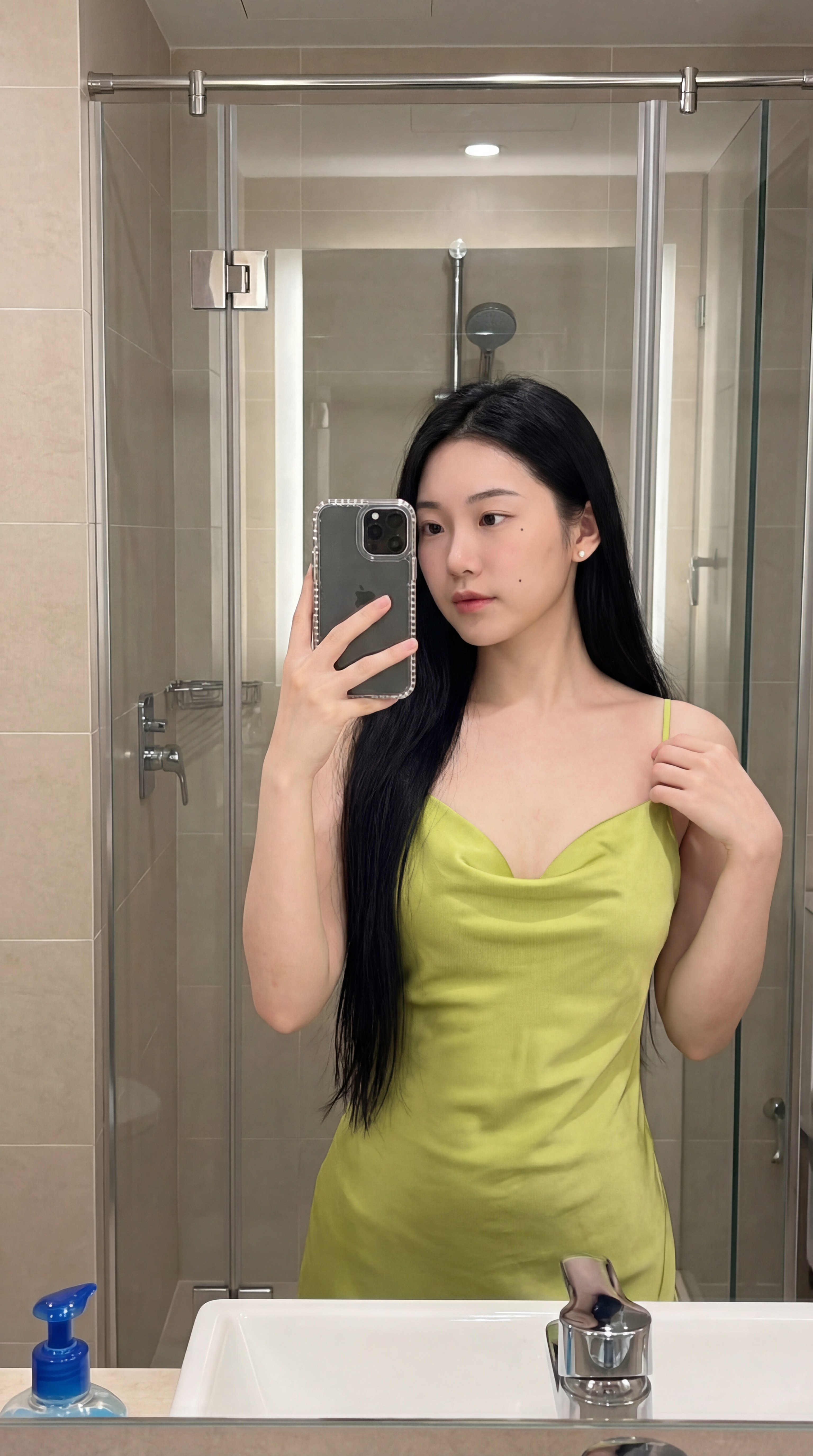 图片展示了一位长发女性在浴室镜子前自拍的场景。她穿着黄色吊带裙，左手扶着镜子，右手拿着手机，手机壳为黑色。背景中可以看到浴室的玻璃门、水龙头、洗手池以及洗手液瓶等物品。该图片是文档中“超简单的日常使用流程”部分的原图，用于说明使用Vision-to-JSON工具时上传图片的示例。