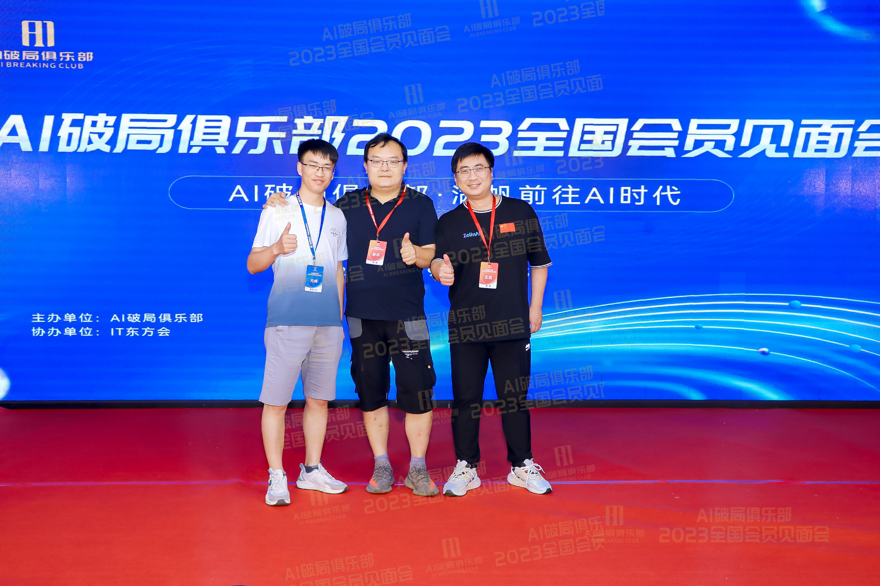 图片展示了AI破局俱乐部2023全国会员见面会的场景。三位男士站在红色地毯上，背景为蓝色带有水波纹的屏幕，屏幕上显示“AI破局俱乐部 2023全国会员见面会”等字样。左侧男士穿浅色短袖和短裤，中间男士穿深色短袖和短裤，右侧男士穿黑色短袖和长裤。他们面带微笑，竖起大拇指，显得十分开心。此图与上下文紧密相关，是对大会中与会人员交流互动场景的呈现。