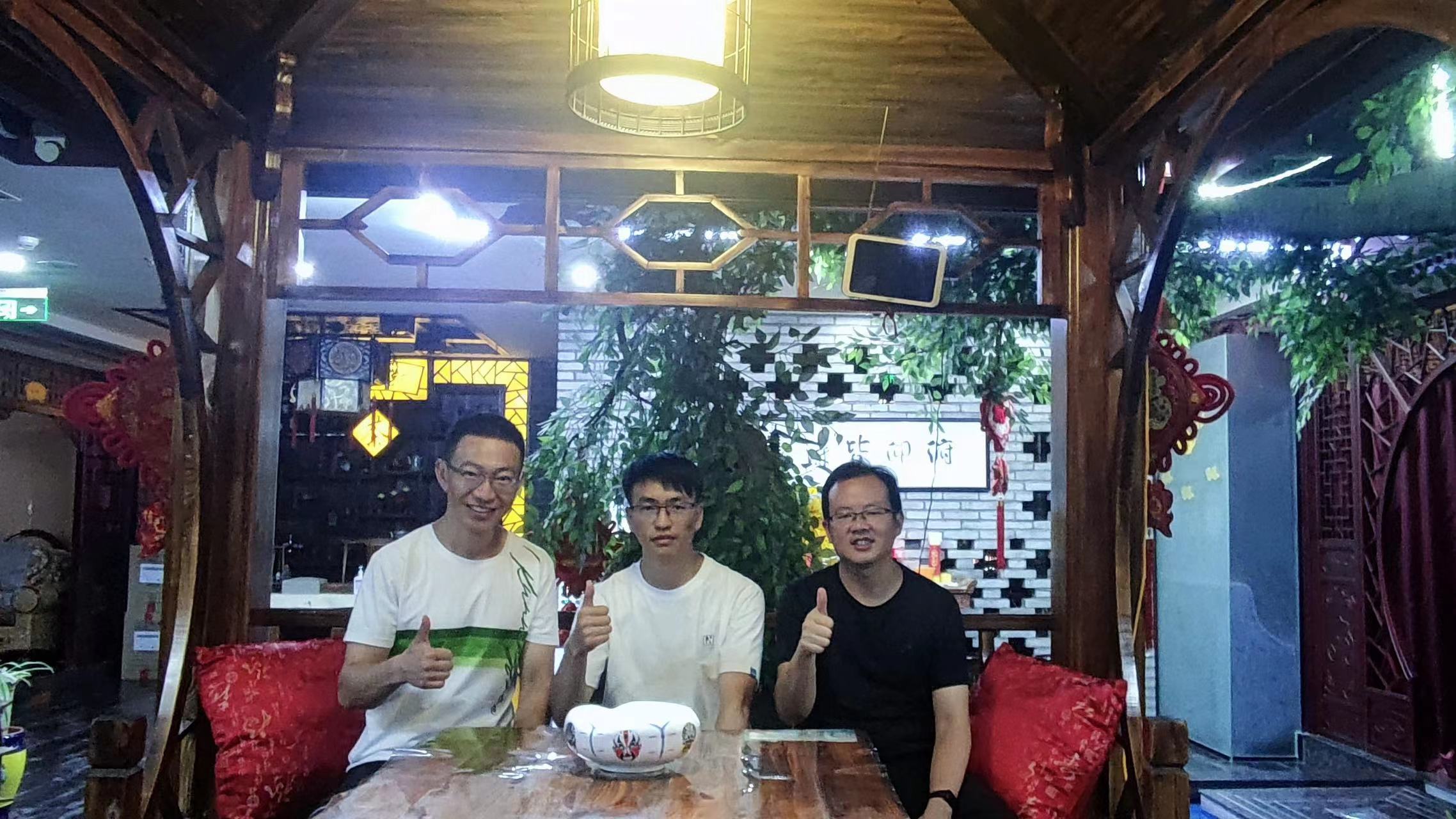 图片展示了三位男士在室内环境中合影。他们坐在一张木桌旁，桌上放着一个白色碗。三人面带微笑，竖起大拇指，显得十分开心。背景为木质结构，有红色靠垫，墙上挂着装饰画，还有一块写有“欢迎光临”的牌匾。整体环境显得温馨且有特色，可能是餐厅或茶馆。此图与上下文关系紧密，可能是记录了与会者在大会期间的友好交流时刻。