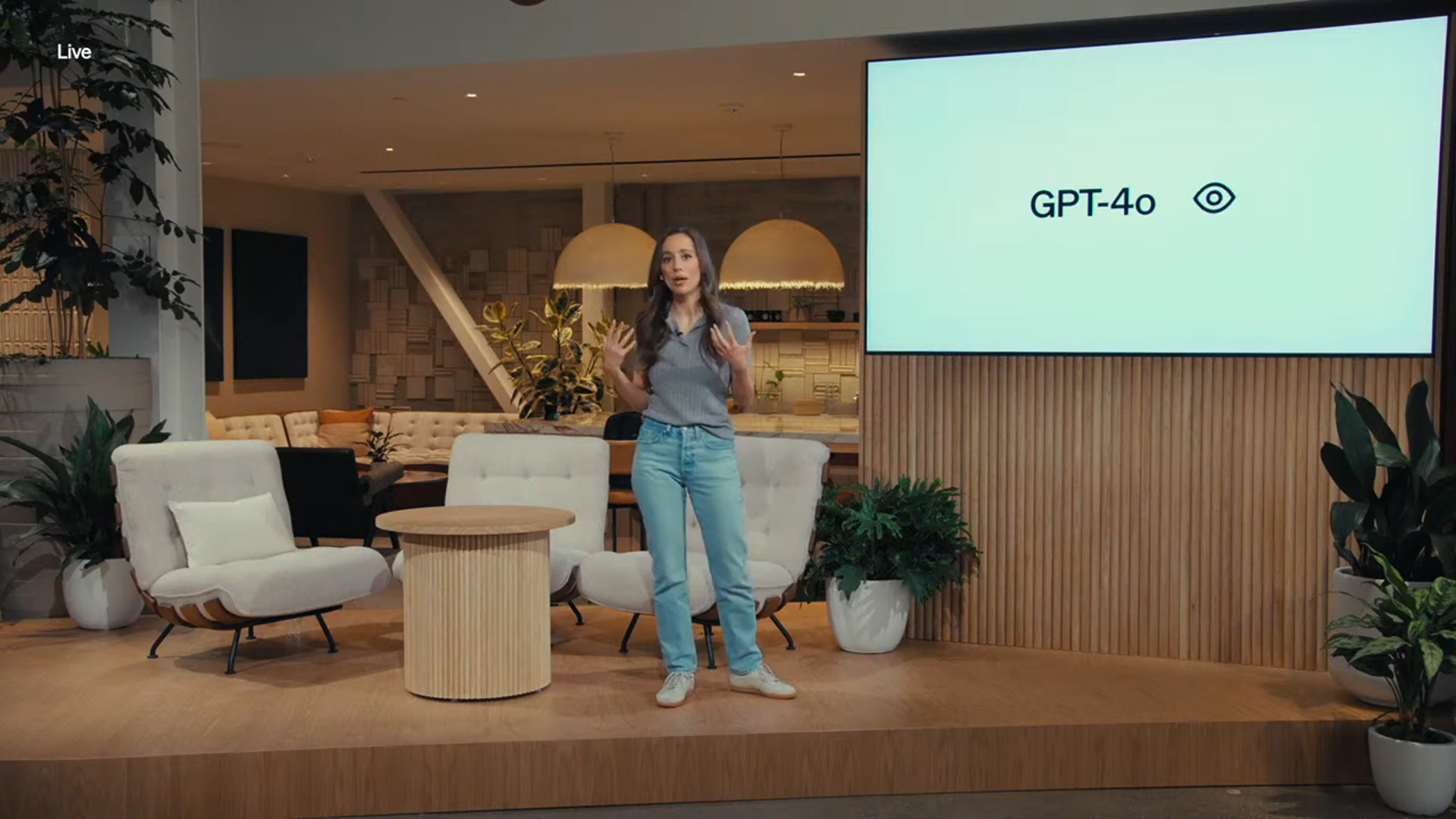 图片展示了一位女性站在一个室内场景中进行讲解，背景屏幕上显示着“GPT-4o”字样及一个眼睛图标。场景布置有舒适的座椅、圆形小桌和绿植，营造出轻松的氛围。左上角有“Live”标识，表明可能是直播场景。这张图片与上下文紧密相关，直观呈现了GPT-4o这一主题，配合文字对GPT-4o的介绍，如模型特性、用户开放范围、价格优势等内容，帮助读者更形象地了解GPT-4o。