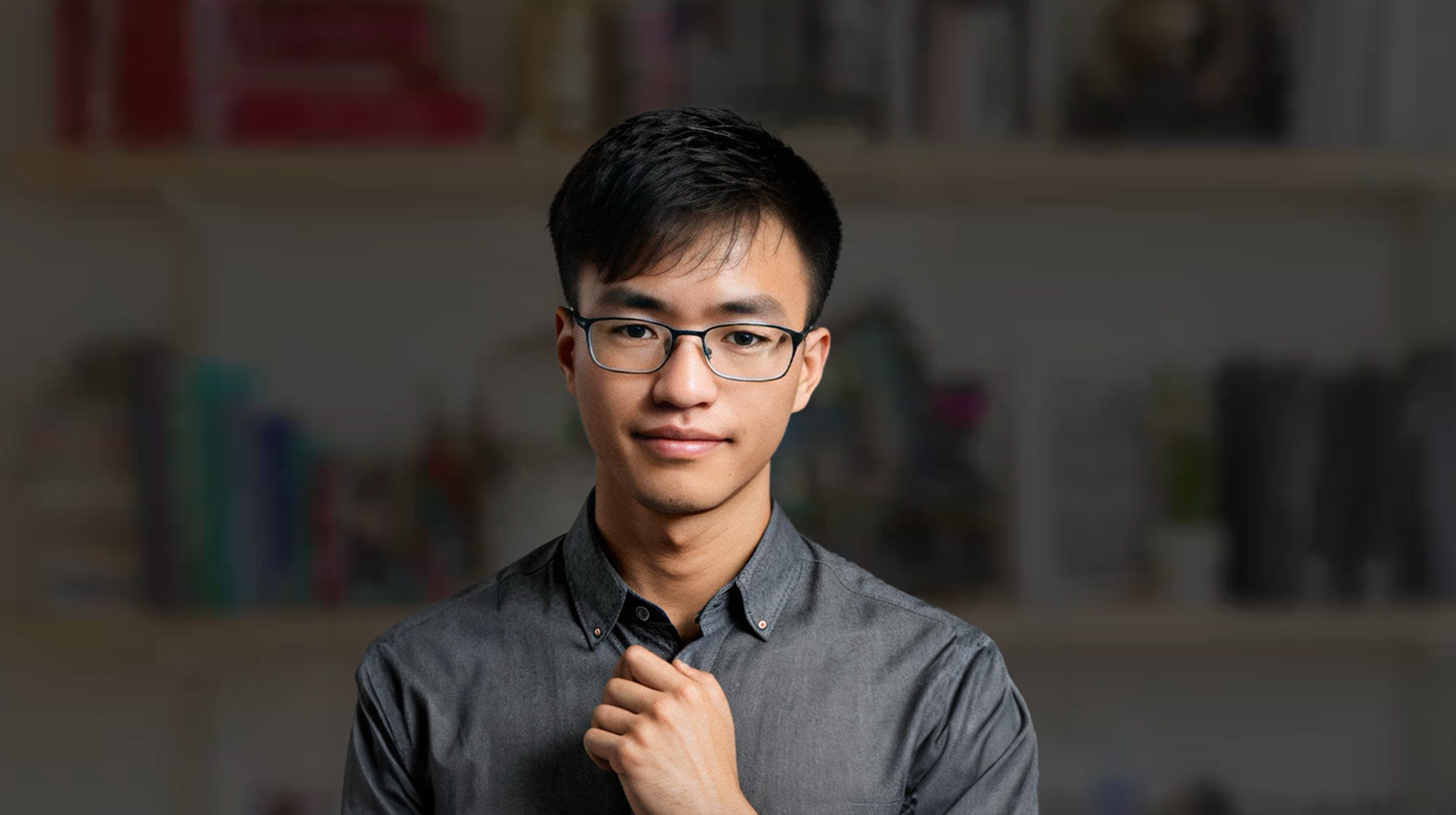 图片展示了一位年轻男子，他戴着眼镜，穿着灰色衬衫，右手轻触衣领。背景为模糊的室内环境，有书架和一些装饰品。该图片位于介绍视频幕后制作内容的文档中，是视频中“我自己的形象”所用的图片，是用Remini写真软件制作的，相比通过Midjourney画要节约很多时间。