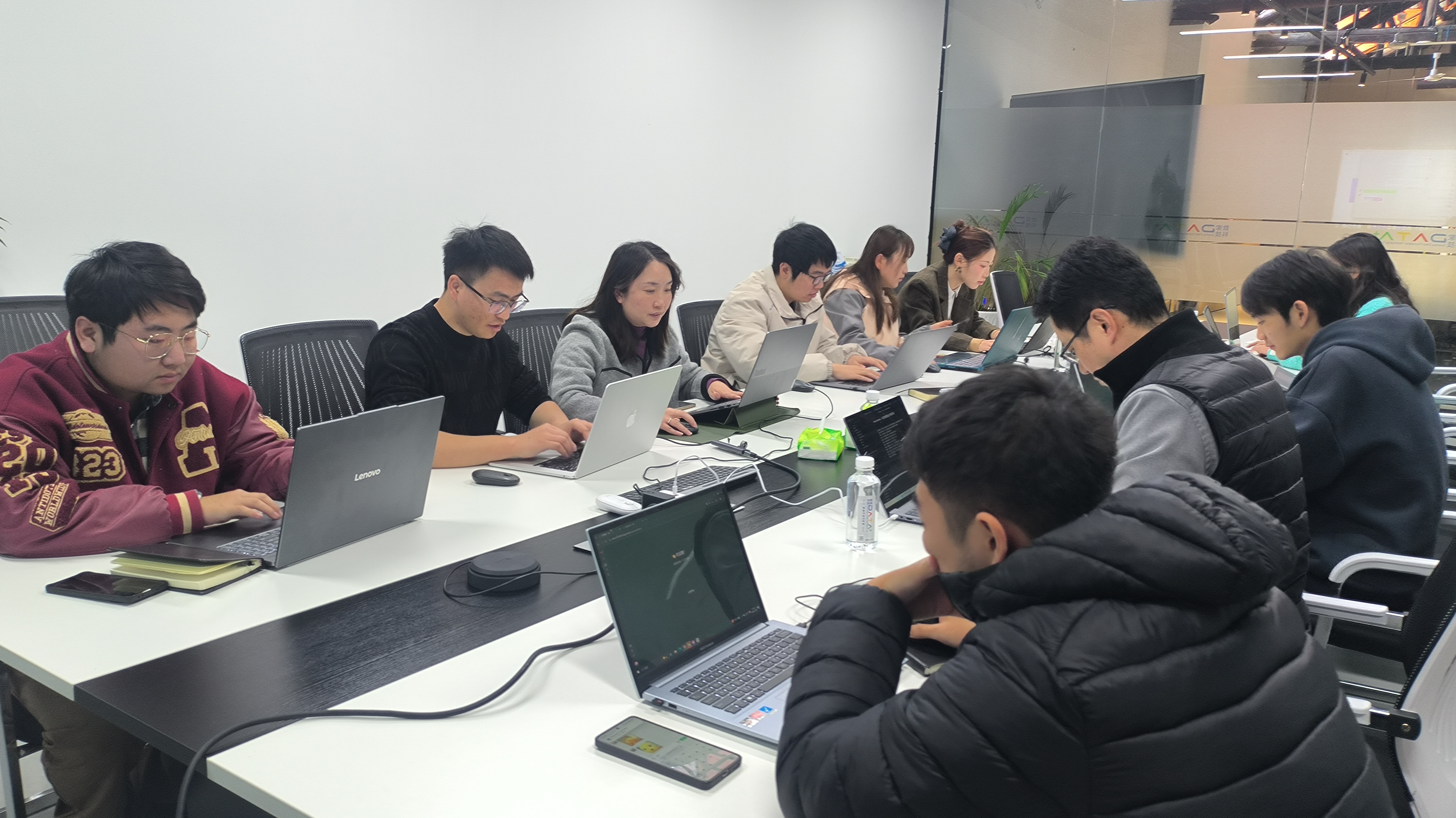 图片展示的是昨天培训现场的场景。多位学员围坐在长桌旁,专注地使用笔记本电脑。桌面上摆放着水瓶、手机等物品。背景中可以看到墙上的装饰和一些植物。这张图片与文档中“昨天培训现场,我发现了小白学N8N的三大死穴”的内容相呼应,直观呈现了培训现场学员学习N8N工作流的场景,为描述小白学N8N时可能遇到的困难提供了直观的背景。