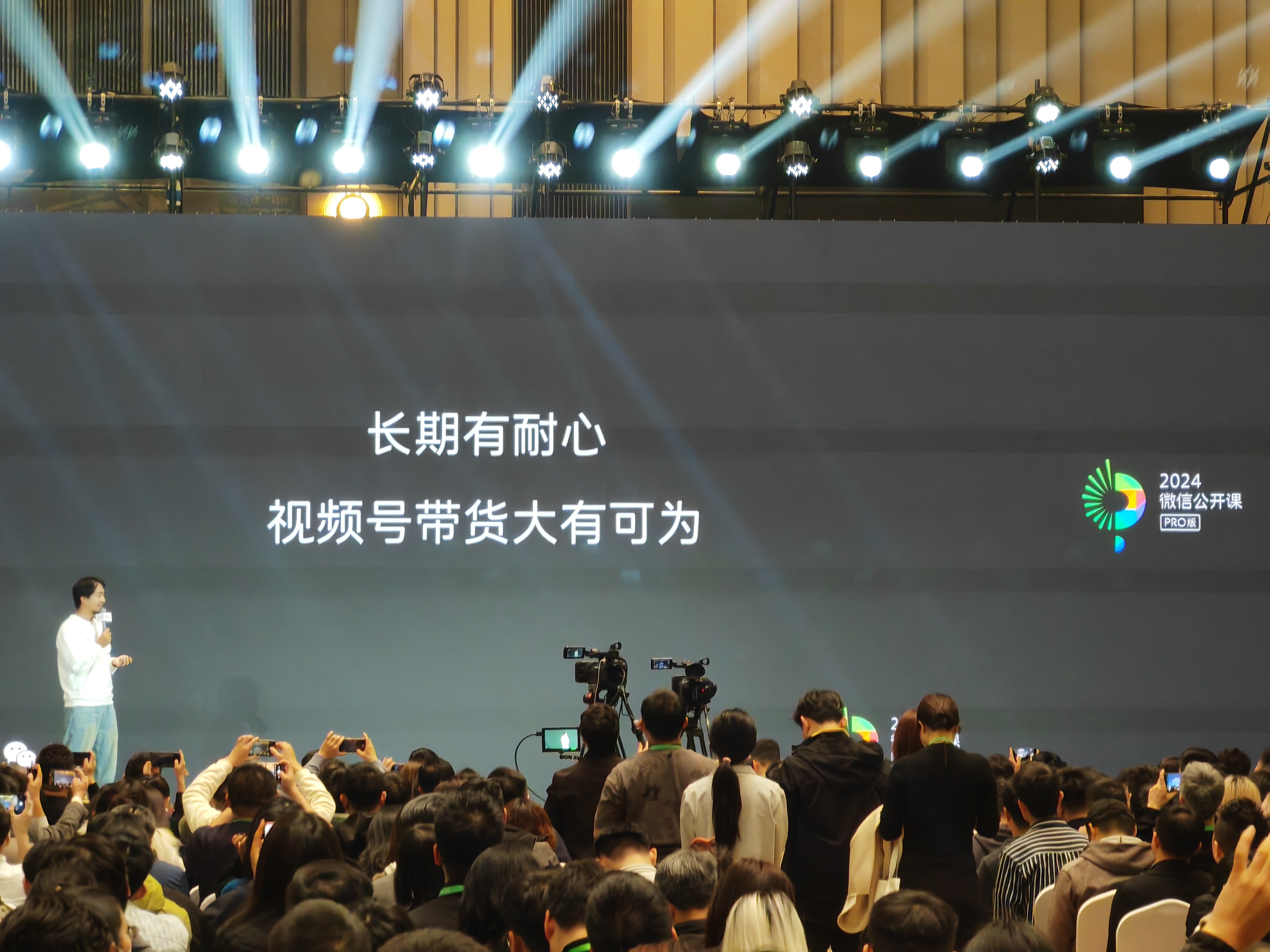 图片展示了2024微信公开课PRO版的现场场景。一位演讲者站在舞台左侧,手持话筒正在讲话。舞台背景大屏上写着“长期有耐心 视频号带货大有可为”,右上角有2024微信公开课PRO版标识。台下聚集着众多观众,部分人举起手机拍照或录像,还有摄像机在进行录制。这张图片与文档中关于2024微信公开课视频号直播带货分享会的内容相呼应,直观呈现了公开课现场氛围。