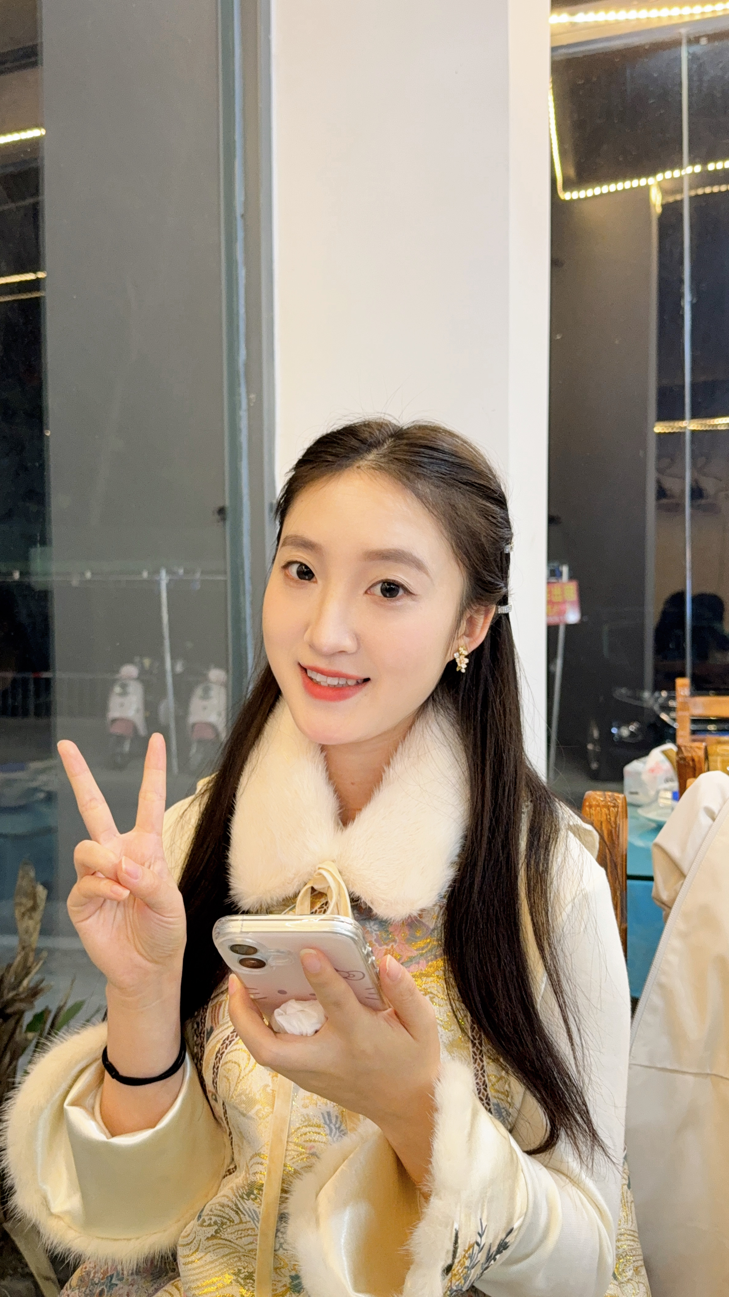 图片展示了一位年轻女性,她面带微笑,右手比出“V”字手势,左手持手机。她穿着白色毛领外套,内搭黄色花纹上衣,耳戴耳环。背景为室内环境,左侧有玻璃隔断,右侧有